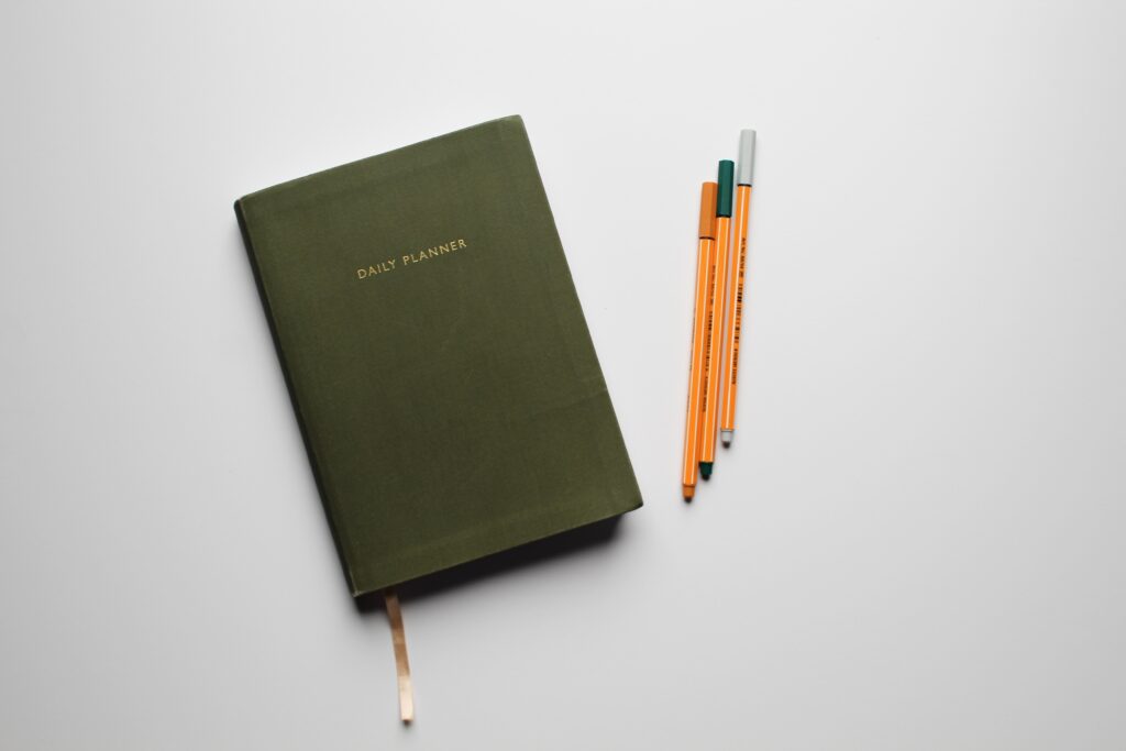 A green book that was used to create a handy travel itinerary can be seen on top of a grey background. Three orange pens are placed to the right of the book.